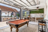 Located at 401 N Main St in the heart of downtown Santa Ana, this rooftop space combines sophisticated indoor and outdoor areas into a cohesive, design-forward venue. Inside, polished terrazzo floors anchor an elegant lounge featuring a dramatic series of arched alcoves draped with flowing white curtains and outfitted with plush, tufted burnt-orange sofas. A striking living moss wall serves as a focal point behind a sleek bar counter with leather-topped stools, while a mosaic tile accent and a projector overhead add both artistry and functionality to the space. Large roll-up garage doors seamlessly connect the interior to the expansive open-air terrace, blurring the line between inside and out. The outdoor area is centered around a wood-slatted pergola strung with bistro lights, beneath which a long communal dining table surrounded by woven rattan chairs creates an inviting gathering space. A built-in outdoor kitchen with dual grills and raised planter beds with lush tropical foliage complete the entertainment zone. A dedicated lawn area with turf and a cornhole game adds a playful touch, while the open perimeter offers sweeping panoramic views of the Santa Ana skyline — making this rooftop an exceptional setting for events or community gatherings.