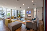 This thoughtfully designed amenity floor offers a dynamic collection of social and recreational spaces united by warm wood-look flooring, bold color accents, and an abundance of natural light pouring in through expansive floor-to-ceiling windows. The main lounge area is a generously scaled gathering space anchored by plush sectional sofas, mustard-yellow accent chairs, and a vibrant multicolored striped feature wall that adds a playful artistic touch. A wall-mounted TV and electric fireplace serve as focal points, while the open layout encourages relaxed conversation and community interaction. Adjacent to the lounge, a well-appointed kitchen and bar area features sleek dark wood cabinetry, a large island with quartz-style countertops, integrated beverage coolers, and pendant lighting — ideal for entertaining. A dedicated media room offers a more intimate retreat, with a large-screen TV set against a teal accent wall, leather seating, and a chevron-patterned rug. The game room makes a bold statement with a full-wall graffiti-style mural and a shuffleboard table. A bright, windowed reading nook with rocking chairs overlooks the outdoors, and a small private meeting room with vibrant artwork and a round table provides a focused workspace. Throughout, curated art, lush greenery, and panoramic views elevate the overall ambiance.