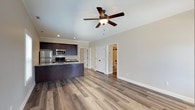 Located at The Cottages, this single-floor residence offers a thoughtfully designed open-concept layout that combines comfort and modern functionality. The kitchen is a standout feature, showcasing rich dark cabinetry with a contrasting granite countertop island, stainless steel appliances including a refrigerator, microwave, and range, making it both practical and visually striking. The kitchen flows naturally into the living area, creating an inviting and connected space ideal for everyday living.

Wide-plank hardwood-style flooring extends throughout the main living area, adding warmth and a clean, contemporary aesthetic. A ceiling fan with a built-in light fixture anchors the living space, providing both comfort and overhead illumination. Neutral wall tones complement the flooring and cabinetry, contributing to the overall bright and airy ambiance.

Crisp white panel doors lead to additional rooms, including a bathroom visible in the background, which features clean, functional finishes. Recessed lighting and well-placed natural light enhance the open feel of the interior. The single-level design ensures easy accessibility and a cohesive flow throughout the entire space, making this cottage a practical and attractive living environment.