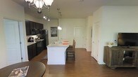 This single-floor space showcases an open-concept layout that seamlessly connects the kitchen and living areas, creating a spacious and cohesive environment. The kitchen is a central highlight, featuring dark espresso cabinetry that contrasts sharply with a white kitchen island equipped with a built-in sink and light-toned countertops. Black appliances, including a refrigerator, microwave, and range, complement the bold cabinetry, lending the kitchen a modern, unified aesthetic. Pendant lighting hangs above the island, providing focused illumination, while a decorative chandelier overhead adds an elegant touch to the dining area nearby. Rich, dark hardwood flooring extends throughout the open space, adding warmth and visual continuity. The neutral, light-toned walls keep the interior feeling bright and airy. Multiple doors visible toward the rear of the space suggest access to additional private rooms and storage areas, including what appears to be a closet with louvered doors. Track lighting on the ceiling supplements the ambient light, ensuring the space is well-lit throughout. Overall, the design balances practicality with style.