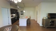 This single-floor space showcases an open-concept layout that seamlessly connects the kitchen and living areas, creating a spacious and cohesive environment. The kitchen is a central highlight, featuring dark espresso cabinetry that contrasts sharply with a white kitchen island equipped with a built-in sink and light-toned countertops. Black appliances, including a refrigerator, microwave, and range, complement the bold cabinetry, lending the kitchen a modern, unified aesthetic. Pendant lighting hangs above the island, providing focused illumination, while a decorative chandelier overhead adds an elegant touch to the dining area nearby. Rich, dark hardwood flooring extends throughout the open space, adding warmth and visual continuity. The neutral, light-toned walls keep the interior feeling bright and airy. Multiple doors visible toward the rear of the space suggest access to additional private rooms and storage areas, including what appears to be a closet with louvered doors. Track lighting on the ceiling supplements the ambient light, ensuring the space is well-lit throughout. Overall, the design balances practicality with style.