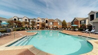 Located at 11010 Eastlake Club Dr in Raleigh, NC, this outdoor amenity area serves as a central gathering space for the community. The focal point is a generously sized, freeform resort-style swimming pool with crystal-clear water, surrounded by a wide brick-paved pool deck lined with lounge chairs and umbrella-shaded seating areas, ideal for relaxing on warm Carolina days.

Adjacent to the pool, a pergola-covered outdoor kitchen and grilling station provides a dedicated space for outdoor cooking and entertaining. The stainless steel grill sits beneath the slatted wood pergola, which frames views of the pool and the surrounding multi-story residential buildings constructed with brick and stucco exteriors.

The expansive brick paver patio extends throughout the area, connecting the pool, outdoor kitchen, and multiple dining and lounge seating arrangements. The space is thoughtfully landscaped with manicured greenery and mature trees that provide natural shade and visual interest.

Access to the amenity area is provided through a glass-paneled teal entry door set into a stucco and brick facade, supported by classical white columns. The overall design blends traditional architectural elements with practical outdoor living features, creating a welcoming and functional community amenity space well-suited to the Raleigh climate.