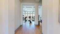 This expansive fitness center offers a comprehensive, multi-zone workout experience spread across a generously sized single-floor layout. The facility is defined by its bright, airy atmosphere, courtesy of large multi-pane windows that flood each room with abundant natural light and provide views of the surrounding outdoors. Soaring ceilings in the main cardio area lend a grand, open feel, complemented by ceiling fans and a coffered ceiling detail that add architectural character.

The cardio zone is home to a full lineup of treadmills, ellipticals, stair climbers, recumbent bikes, and rowing machines, all positioned to take advantage of the panoramic windows. Adjacent strength training areas are outfitted with cable machines, weight racks, dumbbells, benches, and a punching bag, catering to a wide range of workout disciplines. A dedicated cycling and functional training studio features spin bikes, a large wall-mounted TV, floor-to-ceiling mirrors, and plyo boxes for high-intensity training. Throughout the facility, light hardwood-style flooring unifies the space, while white walls and sage-green trim create a clean, welcoming aesthetic. The open entry foyer with structural columns provides a seamless flow between each distinct training area.