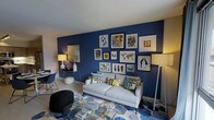 This single-floor, one-bedroom, one-bathroom residence makes excellent use of its thoughtfully arranged layout. The living area serves as the focal point of the home, anchored by a bold navy blue accent wall that creates a striking visual statement. A curated gallery wall displays an eclectic mix of framed artwork in varying sizes, adding personality and artistic depth to the space.

The living room comfortably accommodates a light gray sofa dressed with colorful throw pillows in coordinating hues of blue, gold, and green. A geometric patterned area rug in navy and gray tones grounds the seating area, while a sleek glass coffee table with gold geometric decor accents adds a contemporary touch. A floor lamp and table lamp provide layered ambient lighting throughout the space.

The neutral ceiling and light-toned flooring balance the richness of the navy wall, keeping the space feeling open and bright. A small dining area is visible to the side, featuring modern seating. Overall, the residence presents a cohesive, modern aesthetic with a bold color palette, making it a stylish and functional single-floor living option.