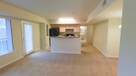This single-floor space is designed with an open-concept layout that integrates the kitchen and living areas into one cohesive environment. The kitchen serves as the functional centerpiece, anchored by a peninsula with a dark stone countertop that provides both workspace and a casual dining or entertaining surface. Light wood cabinetry lines the walls above, complemented by a black refrigerator and an under-cabinet range, while recessed overhead lighting illuminates the cooking area. A pass-through opening connects the kitchen to an adjacent room, likely a dining area or hallway, enhancing the sense of flow throughout the space.

The living area extends generously from the kitchen, finished with neutral-toned wall-to-wall carpeting that adds warmth underfoot. Light-colored walls contribute to a bright, airy atmosphere throughout. A sliding glass door on one side of the living area introduces natural light and provides access to an outdoor space, adding to the overall openness of the layout. The combination of practical finishes, connected living zones, and access to the outdoors makes this a versatile and functional single-floor residence.