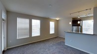 This single-floor residence offers a clean, functional layout with an open-concept design that seamlessly connects the living area to the kitchen. The living space is finished with plush wall-to-wall carpeting in a neutral tone, complemented by soft gray walls that create a calm, inviting atmosphere. Multiple windows fitted with horizontal blinds line the exterior wall, allowing generous amounts of natural light to fill the room while maintaining privacy. The open kitchen is positioned adjacent to the living area and features a raised breakfast bar with granite countertop detailing, providing both a casual dining surface and a natural visual divider between the two spaces. Dark cabinetry overhead contrasts nicely with the lighter countertops, while a stainless steel refrigerator adds a modern, polished touch. A pendant light hangs above the kitchen bar area, offering focused task lighting that enhances the space's functional appeal. The unit's neutral color palette, consistent flooring, and thoughtful open layout make it a versatile and move-in-ready space well-suited for a variety of living arrangements.