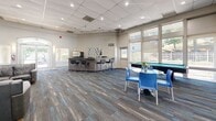 Located at 9584 Muirkirk Rd in Laurel, MD, this combined clubroom and fitness center offers a generously sized, single-floor community amenity space designed for both recreation and exercise. The layout flows seamlessly between distinct functional areas, all unified by stylish multi-toned blue and gray carpet tiles that run throughout.

The fitness center is well-appointed with strength training machines, an elliptical, a stationary bike, and additional cardio equipment, all set against full-length mirrored walls that enhance the sense of space. A bold geometric mural in blue and orange adds an energetic visual focal point. Large floor-to-ceiling windows and a glass sliding door flood the gym with natural light while offering direct views of the outdoor pool area.

The clubroom lounge features comfortable leather sofas, a wall-mounted television, and a round dining table with vibrant blue chairs — creating an inviting space for socializing or relaxing. A kitchenette with a counter-height bar and seating provides a convenient refreshment area, while a billiards table adds a recreational element to the room. Additional windows along the perimeter bring in ample natural light and outdoor views. The space also includes a closet and a private office, rounding out a comprehensive and versatile community facility.