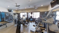 This fitness center offers a thorough and well-organized workout environment designed to accommodate a wide range of exercise needs. The space is outfitted with multiple ceiling fans that promote steady airflow throughout, complementing the recessed lighting overhead for a comfortable and well-lit atmosphere. Large windows along the walls allow natural light to fill the room while providing ventilation, enhancing the overall workout experience.

The cardio section features a row of elliptical machines positioned to take advantage of the natural light from the windows, making for an inviting exercise area. The strength training section is equally comprehensive, housing a multi-station cable and weight machine, a dedicated bench press setup, and a variety of free weights. A collection of stability and medicine balls lines one wall, catering to functional fitness and core training routines.

The flooring throughout is a durable rubber-style surface, ideal for both heavy equipment and high-intensity workouts. The walls are finished in a warm, neutral tone with large mirrors strategically placed to assist with form and to visually expand the space. The layout is thoughtfully arranged to maximize usability while maintaining clear pathways between equipment, creating a safe and efficient training environment.