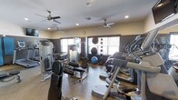 This fitness center offers a thorough and well-organized workout environment designed to accommodate a wide range of exercise needs. The space is outfitted with multiple ceiling fans that promote steady airflow throughout, complementing the recessed lighting overhead for a comfortable and well-lit atmosphere. Large windows along the walls allow natural light to fill the room while providing ventilation, enhancing the overall workout experience.

The cardio section features a row of elliptical machines positioned to take advantage of the natural light from the windows, making for an inviting exercise area. The strength training section is equally comprehensive, housing a multi-station cable and weight machine, a dedicated bench press setup, and a variety of free weights. A collection of stability and medicine balls lines one wall, catering to functional fitness and core training routines.

The flooring throughout is a durable rubber-style surface, ideal for both heavy equipment and high-intensity workouts. The walls are finished in a warm, neutral tone with large mirrors strategically placed to assist with form and to visually expand the space. The layout is thoughtfully arranged to maximize usability while maintaining clear pathways between equipment, creating a safe and efficient training environment.
