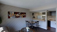 This single-floor residence offers a comfortable and characterful living environment with thoughtfully designed spaces that flow naturally into one another. A standout feature of the dining area is the bold diagonal shiplap accent wall, painted in a soft gray tone, which adds architectural interest and a rustic yet modern charm to the interior. A vibrant abstract canvas artwork adorns the wall, injecting a splash of color and personality into the space.

The dining area comfortably accommodates a round table with ornate wrought-iron chairs, creating an intimate setting well-suited for casual meals or entertaining. A textured popcorn ceiling adds a classic residential character throughout.

An open pass-through counter connects the dining space to the kitchen, promoting an open-concept feel while maintaining defined zones. The kitchen is equipped with standard appliances, including a full-size refrigerator and overhead microwave, and features ample counter space along a wraparound layout. A small decorative plant on the counter adds a touch of greenery.

The flooring transitions from carpet in the dining area to a smooth surface in the kitchen, and the neutral color palette throughout the space creates a cohesive and inviting atmosphere. Small framed photo arrangements on the wall contribute a personal, homey touch to the overall design.