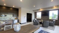This single-floor space offers a well-appointed communal lounge designed with a clean, contemporary aesthetic. The open layout seamlessly connects a compact kitchenette to a comfortable lounge area, creating a functional yet inviting environment. The kitchenette is outfitted with dark cabinetry, a tile backsplash, and a mini-refrigerator, providing convenient amenities for residents or guests. Recessed lighting throughout the ceiling ensures consistent, even illumination across the space.

The lounge area features a grouping of gray upholstered armchairs arranged around a wooden coffee table, encouraging conversation and relaxation. An exposed brick accent wall adds warmth and texture, contrasting tastefully with the smooth white walls and light-toned flooring. A wall-mounted flat-screen television is positioned above the brick feature, enhancing the space's entertainment value. A window provides a glimpse of natural light and outdoor views, brightening the seating area. A rustic wooden table in the foreground, accented with decorative lanterns, adds a homey touch to the otherwise modern setting.