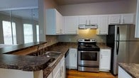 This single-floor residence offers a thoughtfully designed layout centered around a fully equipped kitchen. The kitchen stands out with its crisp white cabinetry providing ample storage, dark granite-style countertops that add a rich contrast, and warm hardwood flooring that runs throughout. A stainless steel double-basin sink is positioned beneath a window, allowing natural light to fill the space, while a stainless steel electric range with an overhead ventilation hood serves as the functional centerpiece for cooking. The kitchen's L-shaped configuration maximizes workspace and flow, making it both practical and visually appealing. The neutral wall tones complement the cabinetry and countertops, creating a cohesive and inviting atmosphere. A dishwasher is seamlessly integrated beneath the counter, contributing to the kitchen's clean, streamlined aesthetic. The overall ambiance of the space is warm and welcoming, with a balance of classic design elements and modern appliances that make it well-suited for everyday living and entertaining alike.
