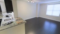 This single-floor space showcases a thoughtfully designed open-concept layout that seamlessly connects the kitchen and living areas. Dark hardwood flooring runs throughout, providing a rich contrast against the bright white walls and crisp white baseboards, creating a clean and polished aesthetic.

Crown molding lines the ceiling, adding an elegant architectural detail that elevates the overall finish of the space. A track lighting fixture is mounted on the ceiling, providing focused, adjustable illumination across the open living area. A window along the far wall allows for natural light to filter into the space.

The kitchen features a granite countertop peninsula with an overhang that accommodates seating, making it a practical and social focal point. The granite surface displays a neutral speckled pattern that complements the light cabinetry below. The peninsula serves as a natural visual divider between the kitchen and the open living space without disrupting the airy, open-concept feel.

Overall, the space presents a bright, clean atmosphere with consistent finishes, including coordinated flooring, trim work, and countertops, making it a well-appointed and cohesive single-level living environment.