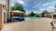 Located at University Suites in Greenville, North Carolina, this outdoor amenity space offers a generously sized swimming pool as its centerpiece, surrounded by a well-maintained concrete sundeck that provides ample room for lounging and socializing. The pool features clearly marked depth indicators and easy-access entry points, making it both functional and safe for residents.

A standout feature of the space is the wooden pergola-style cabana situated adjacent to the pool deck. Outfitted with comfortable seating upholstered in vibrant teal cushions, the covered lounge area offers a shaded retreat from the sun while maintaining an open, airy feel. Additional seating is provided throughout the deck in the form of multiple chaise lounges arranged along the pool's perimeter, ideal for sunbathing.

The space is framed by mature trees that provide a natural privacy screen and a lush green backdrop, enhancing the resort-like atmosphere. A brick building with a pitched roof serves as a poolside facility structure, likely housing restrooms or equipment, and integrates seamlessly into the overall aesthetic. A wrought-iron perimeter fence encloses the area, ensuring security while maintaining an open sightline to the surrounding landscape. The entire space is thoughtfully designed to support outdoor recreation and relaxation.
