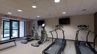 This fitness room offers a focused exercise environment outfitted with a selection of cardio equipment, including a treadmill and two elliptical machines. The space is designed for efficient use, with each machine positioned to allow comfortable movement and access. Two wall-mounted flat-screen televisions are strategically placed to keep users entertained during workouts. The room features a drop ceiling with recessed circular lighting fixtures that provide even, functional illumination throughout. The walls are finished with a neutral, textured wallcovering that contributes to a clean and professional atmosphere. A large window along one side of the room introduces natural light, adding an element of openness to the space. The flooring is light-toned and easy to maintain, complementing the overall utilitarian yet welcoming design. The layout is straightforward and uncluttered, prioritizing practicality and ease of use. Overall, this fitness room provides a well-equipped and comfortable setting for cardiovascular exercise within a single-level facility.