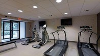 This fitness room offers a focused exercise environment outfitted with a selection of cardio equipment, including a treadmill and two elliptical machines. The space is designed for efficient use, with each machine positioned to allow comfortable movement and access. Two wall-mounted flat-screen televisions are strategically placed to keep users entertained during workouts. The room features a drop ceiling with recessed circular lighting fixtures that provide even, functional illumination throughout. The walls are finished with a neutral, textured wallcovering that contributes to a clean and professional atmosphere. A large window along one side of the room introduces natural light, adding an element of openness to the space. The flooring is light-toned and easy to maintain, complementing the overall utilitarian yet welcoming design. The layout is straightforward and uncluttered, prioritizing practicality and ease of use. Overall, this fitness room provides a well-equipped and comfortable setting for cardiovascular exercise within a single-level facility.