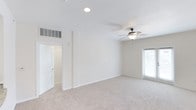 This single-floor space presents a clean, well-maintained interior centered around a generously sized main room. The room is finished with soft, neutral-toned carpet that extends throughout, creating a warm and comfortable underfoot experience. Walls are painted in a light beige tone, contributing to an open and airy atmosphere.

Overhead, a dark bronze ceiling fan with a built-in light fixture provides both ventilation and supplemental lighting, complementing a recessed ceiling light that ensures the space is well-illuminated. A window fitted with horizontal blinds along one wall allows natural light to filter into the room while offering privacy control.

A clearly visible HVAC vent is positioned high on the wall, indicating a centralized climate control system. White panel doors lead to additional areas of the unit, suggesting connectivity to other rooms or storage spaces. Clean white baseboards frame the perimeter of the room, adding a crisp contrast to the neutral walls and carpet. Overall, the space offers a practical, move-in-ready environment with a simple, functional aesthetic suited to a variety of uses.
