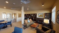 This single-floor communal lounge space is thoughtfully designed to accommodate a variety of social and leisure activities. The main area is anchored by a fireplace flanked by dark wood built-in bookshelves filled with books and decorative accents, creating a warm and inviting focal point. A comfortable sofa with accent pillows and a leather-topped coffee table complement the seating arrangement, while additional blue upholstered chairs and round wood-top tables are arranged throughout to encourage both casual conversation and small group gatherings. Recessed lighting and a central ceiling-mounted HVAC unit keep the space well-lit and climate-controlled. Neutral carpet flooring and warm beige walls lend a calm, welcoming ambiance throughout. Large windows along one side of the space allow natural light to pour in, offering views of the surrounding exterior. Framed landscape artwork adorns the walls, adding visual interest and a sense of place. A pass-through opening connects to an adjacent area, while a barn-style glass door leads to a private meeting or office room equipped with its own workspace. Additional shelving and seating in this ancillary space make it ideal for quieter, more focused activities. Overall, the layout balances open communal areas with more intimate zones, making it a versatile and functional shared space.