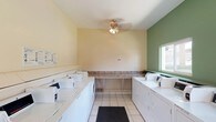 This single-story shared laundry facility is designed for functionality and efficiency, offering a well-organized layout that accommodates multiple users simultaneously. Rows of commercial-grade top-load washers and dryers line both sides of the room, providing ample capacity for high-volume use. The machines are equipped with digital control panels, and posted instructional signage assists users with proper operation.

A continuous laminate folding counter runs along one wall, offering generous workspace for sorting and folding laundry. The room features white ceramic tile flooring that is easy to maintain, complemented by cream-colored walls and a bold green accent wall that adds a touch of visual interest. A ceiling fan is centrally mounted overhead, promoting air circulation throughout the space.

Natural light enters through a side window and a glazed entry door, creating a bright and welcoming atmosphere. The entry door also provides a direct outdoor view, enhancing ventilation options. A stainless steel waste bin is conveniently positioned near the entrance. A no-smoking sign is prominently displayed, reinforcing the facility's usage policies. Overall, the space is clean, practical, and well-equipped for communal laundry needs.