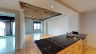 This single-floor space showcases an industrial-modern aesthetic defined by its exposed concrete ceiling, which serves as a striking architectural focal point. An open-plan layout seamlessly connects the kitchen and living areas, creating a spacious, versatile environment. The kitchen features rich, dark granite countertops with visible mineral detailing, warm wood cabinetry with brushed metal hardware, and a stainless steel sink with a modern arc faucet. Overhead, track lighting mounted to the concrete ceiling provides adjustable illumination throughout the space, complementing the recessed lighting integrated into the dropped ceiling section. A large window at the far end of the living area allows natural light to filter in, brightening the white walls and enhancing the reflective quality of the light hardwood floors. An exposed cylindrical ventilation duct runs along the ceiling, adding to the loft-inspired industrial character of the space. The clean white walls throughout create a neutral backdrop that balances the rawness of the exposed structural elements, resulting in a space that is both functional and architecturally compelling.