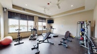 Located at 1410 Deer Lakes Dr in Buffalo, NY, this dedicated exercise room offers a well-appointed fitness facility designed to support a wide range of workout routines. The space features durable rubber flooring throughout, providing a safe and functional surface for exercise. A tray ceiling with recessed lighting lends a polished architectural touch, while a ceiling fan ensures comfortable air circulation during workouts.

Natural light floods the room through a series of large windows dressed with dark blinds, offering pleasant views of the surrounding landscape and water, creating an inspiring backdrop for exercise. A large wall-mounted mirror spans one side of the room, visually expanding the space and aiding in form correction during training.

The room is outfitted with a diverse selection of equipment, including treadmills and an elliptical machine from the TRUE brand, stationary and spin bikes, a cable/pulley strength training machine, an adjustable workout bench, a dumbbell rack, medicine balls, and stability balls. A wall-mounted flat-screen television adds entertainment options for users. The space also includes a designated entry door and informational postings, reflecting an organized, community-oriented fitness environment.