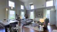 Located at 4115 Britton Parkway in Hilliard, Ohio, this single-story space makes an immediate impression with its dramatic vaulted ceilings that rise to striking heights, creating a sense of openness and grandeur. The main living area is bathed in natural light, courtesy of a generous arrangement of tall windows positioned along multiple walls, as well as a skylight that draws sunlight deep into the interior throughout the day. Rich hardwood flooring runs throughout, grounding the space with warmth and texture. The living area is thoughtfully appointed, featuring a neutral gray color palette on the walls that complements the contemporary furnishings. A large statement clock serves as a bold decorative focal point, while built-in and freestanding shelving units offer both style and function. The open layout seamlessly integrates a sitting and workspace area, making the space highly versatile. Sheer, patterned drapery frames the many windows, softening the abundant natural light while adding an elegant touch. The overall ambiance is modern and inviting, balancing clean architectural lines with carefully curated décor. This well-designed space offers a bright, comfortable, and functionally flexible environment in a desirable Hilliard location.