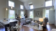 Located at 4115 Britton Parkway in Hilliard, Ohio, this single-story space makes an immediate impression with its dramatic vaulted ceilings that rise to striking heights, creating a sense of openness and grandeur. The main living area is bathed in natural light, courtesy of a generous arrangement of tall windows positioned along multiple walls, as well as a skylight that draws sunlight deep into the interior throughout the day. Rich hardwood flooring runs throughout, grounding the space with warmth and texture. The living area is thoughtfully appointed, featuring a neutral gray color palette on the walls that complements the contemporary furnishings. A large statement clock serves as a bold decorative focal point, while built-in and freestanding shelving units offer both style and function. The open layout seamlessly integrates a sitting and workspace area, making the space highly versatile. Sheer, patterned drapery frames the many windows, softening the abundant natural light while adding an elegant touch. The overall ambiance is modern and inviting, balancing clean architectural lines with carefully curated décor. This well-designed space offers a bright, comfortable, and functionally flexible environment in a desirable Hilliard location.