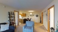 This single-floor residence offers a well-organized and inviting layout with an open-concept design that connects the main living spaces seamlessly. The living area features plush carpeting and is accented by a decorative pendant light fixture that adds a warm, homey ambiance. A leaning bookshelf near the entryway provides both functional storage and a personal touch.

The kitchen is equipped with warm honey-toned wood cabinetry, a microwave, and ample counter space, making it both practical and visually cohesive. Adjacent to the kitchen, the dining area accommodates a full table and chairs, making it ideal for everyday meals or casual gatherings.

The main entry door features a charming decorative wreath, contributing to the welcoming atmosphere of the home. Additional doors throughout the space lead to separate rooms, suggesting a thoughtful and functional floor plan. The neutral color palette of the walls and flooring creates a bright, clean backdrop that complements the warm wood tones and patterned soft furnishings visible throughout. Overall, this residence offers a comfortable and efficiently designed living environment suited for everyday living.