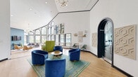Located at 2 Wyndham Dr in Hampton, Virginia, this single-floor community space offers a thoughtfully designed, multi-functional interior that blends modern aesthetics with a welcoming atmosphere. The space is unified by warm hardwood-style flooring and crisp white walls accented with bold navy trim, creating a clean and contemporary feel throughout.

The living area is defined by dramatic, floor-to-ceiling arched windows that flood the interior with abundant natural light and frame views of the surrounding greenery. Soaring ceilings further amplify the sense of openness, while a striking multi-globe chandelier adds an elegant focal point. This lounge zone is furnished with a mix of tan leather accent chairs and a plush grey sofa, arranged around patterned area rugs for a relaxed yet refined setting.

A dedicated workspace with a built-in desk and workstation sits adjacent to the main lounge, providing a functional area for residents. A separate seating nook features rich cobalt blue velvet chairs grouped around a round coffee table, anchored beneath an arched interior doorway that adds architectural charm.

The refreshment bar area showcases a bold hexagonal mosaic tile backsplash, teal cabinetry, and a butcher block countertop with bar seating — a vibrant and stylish amenity. Double glass-panel entry doors complete this inviting, well-appointed community hub.