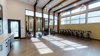 This contemporary fitness studio is designed to inspire and energize, combining stylish architectural details with a fully equipped workout environment. The space features soaring ceilings accented by rich dark wood beams and elegant globe chandeliers, creating an atmosphere that balances functionality with refined design. Large floor-to-ceiling windows line the walls, flooding the hardwood floors with generous natural light and offering a bright, open ambiance throughout the day. Full-length mirrors along one wall enhance the sense of space while providing essential feedback for form and technique during workouts. The studio is well-appointed with an array of fitness equipment, including a row of stationary cycling bikes along one side, perfect for high-intensity cardio sessions. A dedicated free weights and functional training area features medicine balls, BOSU balance trainers, and a rack of dumbbells, catering to strength and conditioning needs. The open floor plan allows for versatile use of the space, accommodating group fitness classes, personal training, or individual workouts with ease. The warm neutral-toned walls and natural wood flooring contribute to a welcoming and motivating environment, making this fitness studio a standout facility for health and wellness.