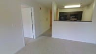 This single-floor residential unit features an open, functional layout that connects the living area to the kitchen through a half-wall pass-through counter. The living space is finished with light-toned wall-to-wall carpeting, lending a clean and neutral aesthetic throughout. The kitchen is positioned at an elevated counter height relative to the living area, providing a natural visual separation while maintaining an open feel. It is equipped with dark cabinetry, a black refrigerator, and a built-in microwave, with overhead fluorescent lighting illuminating the workspace. A doorway visible from the living area leads to a hallway, suggesting additional rooms beyond the main living space. The walls are painted in a soft, neutral tone that complements the light carpeting. A wall vent and a fire alarm are visible, reflecting standard residential utility installations. The overall layout is straightforward and practical, with clearly defined zones for living and cooking. The design prioritizes ease of movement and functional use of space, making it a practical residential setting with an accessible, single-level floor plan.