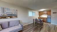 This single-floor unit offers an efficiently arranged layout that maximizes every inch of its compact footprint. Consistent wood-style flooring flows throughout the entire space, lending a cohesive and modern feel. The textured ceilings add subtle architectural character to each room.

The open-plan main area seamlessly integrates the living room, dining area, and kitchen. The living room provides a comfortable lounging space, brightened by windows fitted with vertical blinds that diffuse natural light softly across the room. A wall-mounted PTAC unit ensures climate control year-round. The dining area accommodates a modest dining set positioned adjacent to a large window overlooking outdoor greenery. The kitchen features warm-toned wood cabinetry, granite-style countertops, a stainless steel sink, a built-in cooktop, a stainless steel refrigerator, and an overhead range hood — offering practical functionality in a compact arrangement.

The bedroom is a well-proportioned private retreat, comfortably accommodating a full-sized bed alongside additional furnishings. A large window with vertical blinds allows natural light while providing privacy. A baseboard heater along the wall ensures warmth. Overall, the unit presents a clean, functional, and inviting atmosphere.