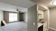 This single-floor residence offers a practical and comfortable layout designed for everyday living. The living area is welcoming and open, finished with plush carpeting and neutral gray-toned walls that create a calm, cohesive atmosphere. A ceiling fan with a built-in light fixture provides both comfort and illumination, while a large window allows natural light to fill the space and offers views to the outdoors. The kitchen is a standout feature, configured as a galley-style corridor with crisp white shaker-style cabinetry, light granite-look countertops, and black appliances including a refrigerator, range, and built-in microwave. Durable wood-look flooring runs the length of the kitchen, contrasting neatly with the carpeted living spaces. Textured walls throughout add visual depth and character to the interior. The additional rooms are generously sized, each with soft carpeting, ceiling fans, and windows that invite natural light. A utility area houses the electrical panel, and the overall layout connects rooms through a central hallway, ensuring an efficient flow across the floor plan. The consistent use of warm neutral paint tones, white trim, and modern fixtures gives the space a clean, move-in-ready feel throughout.