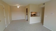 This single-floor residence offers a practical and welcoming layout designed for comfortable everyday living. The main living area is spacious and open, featuring neutral-toned carpet flooring and warm beige walls that create a calm, inviting atmosphere. A ceiling fan provides both style and function overhead, contributing to the room's airy feel.

A defining architectural feature of the space is the kitchen pass-through window, which opens from the kitchen into the living area, creating a sense of connectivity between the two spaces. The kitchen is equipped with under-cabinet lighting that illuminates a granite-style countertop, lending a polished look to the cooking space. Dark wood cabinetry adds warmth and contrast to the kitchen's overall design.

Adjacent to the kitchen, a dedicated laundry area is visible, housing an in-unit washer — a highly practical convenience. The transition between the carpeted living area and the harder flooring of the kitchen and utility spaces is clearly defined, adding visual interest to the layout.

Throughout the space, clean white baseboards frame the rooms neatly, and the neutral color palette ensures versatility for a variety of interior styles. The overall ambiance is understated and functional, with thoughtful design details that enhance both the utility and comfort of daily living.