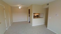 This single-floor residence offers a practical and welcoming layout designed for comfortable everyday living. The main living area is spacious and open, featuring neutral-toned carpet flooring and warm beige walls that create a calm, inviting atmosphere. A ceiling fan provides both style and function overhead, contributing to the room's airy feel.

A defining architectural feature of the space is the kitchen pass-through window, which opens from the kitchen into the living area, creating a sense of connectivity between the two spaces. The kitchen is equipped with under-cabinet lighting that illuminates a granite-style countertop, lending a polished look to the cooking space. Dark wood cabinetry adds warmth and contrast to the kitchen's overall design.

Adjacent to the kitchen, a dedicated laundry area is visible, housing an in-unit washer — a highly practical convenience. The transition between the carpeted living area and the harder flooring of the kitchen and utility spaces is clearly defined, adding visual interest to the layout.

Throughout the space, clean white baseboards frame the rooms neatly, and the neutral color palette ensures versatility for a variety of interior styles. The overall ambiance is understated and functional, with thoughtful design details that enhance both the utility and comfort of daily living.
