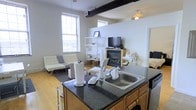 This single-floor space offers an open-concept layout that seamlessly connects the kitchen and living area, making efficient use of the available footprint. The kitchen anchors the space with a central island topped with dark granite-style countertops, a stainless steel double sink, and warm wood cabinetry underneath, providing ample storage and workspace. The open-plan design allows for an unobstructed sightline into the living area, which features light hardwood flooring throughout. An exposed black industrial duct runs along the upper wall, lending the space a loft-like character that is complemented by tall ceilings and large multi-pane windows that invite generous amounts of natural light. The living area is centered around a fireplace, which serves as a focal point and adds warmth to the overall ambiance. A shelving unit provides display and storage space alongside the entertainment setup. The neutral white walls and bright, airy atmosphere create a clean and versatile backdrop. Overall, the space balances functional, practical design with distinctive industrial and cozy touches.