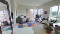 This single-floor living space is designed with a contemporary aesthetic that balances bold color and clean architectural lines. The living area serves as the focal point, featuring large corner windows that flood the room with natural light while framing views of a neighboring brick building and surrounding rooftops. The white walls and light flooring provide a neutral backdrop that allows the colorful furnishings to stand out prominently.

A vibrant patchwork area rug in shades of blue, orange, green, and purple anchors the seating arrangement, which includes a gray upholstered sofa accented with teal and patterned throw pillows, alongside a pair of classic armchairs with navy cushions. A rounded metallic accent table serves as a coffee table, adding an eclectic, modern touch. Colorful abstract artwork adorns the walls, contributing to the room's energetic and curated ambiance.

The space is thoughtfully layered with decorative elements, including potted plants near the windows and mirrored accent furniture that enhances the sense of openness. Overall, the space presents a lively, well-lit, and stylishly furnished interior suited for comfortable everyday living.