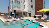 The pool area at South 46 is a well-appointed outdoor amenity space designed for relaxation and leisure. The centerpiece is a vibrant aqua-tiled pool, distinguished by built-in in-water lounge chairs that allow residents to recline directly in the water. The surrounding deck provides ample space for additional seating, including contemporary sling-back chairs arranged for both sun exposure and shade. A covered lounge area along the building's base offers a shaded retreat, furnished with comfortable seating and accented by a grid-style decorative wall feature. The space is framed by the building's modern façade, which combines white paneling with brick exterior elements and floor-to-ceiling windows on the upper floors. Interior corridors connecting to the pool area are finished with patterned gray carpet tiles and teal accent walls, creating a cohesive, modern aesthetic throughout. Wall sconces provide consistent lighting along the hallways, and access doors with exterior views open to a sun-drenched deck overlooking the surrounding landscape. The overall design balances functional amenities with a contemporary, community-focused atmosphere.