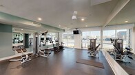 This fitness center offers a comprehensive workout environment spread across a single, thoughtfully organized floor. The space is divided into distinct functional zones, each designed to support a variety of training styles. The main exercise area features large, floor-to-ceiling windows that flood the room with natural light and provide open views of the surrounding exterior, creating an energizing atmosphere. Ceiling fans and recessed lighting contribute to a comfortable workout environment throughout.

The cardio zone houses stationary bikes equipped with digital displays, along with treadmills and elliptical machines visible through a glass-paneled interior wall, offering visual connectivity between areas while maintaining distinct spaces. A dedicated strength training section features a multi-station cable machine and a weight bench, complemented by full-length wall mirrors that enhance the sense of space.

The free weights area is stocked with a tiered dumbbell rack offering a wide range of weights, a weight bench, and stability and medicine balls on a dedicated rack — catering to both beginners and advanced users. Rubber flooring throughout provides safety and comfort underfoot. A generous open stretch of floor allows ample room for mat-based exercises and stretching. The facility is accessed through double glass doors and maintains a clean, modern aesthetic with soft sage-green accent walls and crisp white trim.