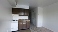 Located at 300 W 3rd Street in Bridgeport, PA 19405, this single-floor space offers a straightforward and functional layout. The kitchen is equipped with dark brown cabinetry, both upper and lower, providing ample storage. A built-in electric range and cooktop sits adjacent to a single-basin sink with matching lower cabinetry underneath. A range hood is mounted above the stove, and a laminate countertop runs the length of the cooking and prep area. The kitchen opens directly into the living area, which features wall-to-wall carpeting and neutral-toned walls, creating a cohesive and open feel. A louvered double-door closet is visible just off the main living space, offering convenient built-in storage. The ceilings are standard height and the walls are painted in a clean, light neutral tone throughout. The open-plan arrangement between the kitchen and living area allows for flexible use of the space. Overall, the unit presents a practical and utilitarian design suitable for a variety of occupants looking for a no-frills, move-in-ready space in the Bridgeport area.