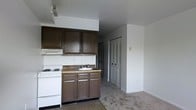 Located at 300 W 3rd Street in Bridgeport, PA 19405, this single-floor space offers a straightforward and functional layout. The kitchen is equipped with dark brown cabinetry, both upper and lower, providing ample storage. A built-in electric range and cooktop sits adjacent to a single-basin sink with matching lower cabinetry underneath. A range hood is mounted above the stove, and a laminate countertop runs the length of the cooking and prep area. The kitchen opens directly into the living area, which features wall-to-wall carpeting and neutral-toned walls, creating a cohesive and open feel. A louvered double-door closet is visible just off the main living space, offering convenient built-in storage. The ceilings are standard height and the walls are painted in a clean, light neutral tone throughout. The open-plan arrangement between the kitchen and living area allows for flexible use of the space. Overall, the unit presents a practical and utilitarian design suitable for a variety of occupants looking for a no-frills, move-in-ready space in the Bridgeport area.