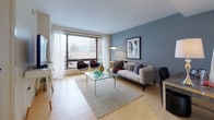 This 1-bedroom, 1-bathroom urban residence is thoughtfully designed with a clean, contemporary aesthetic throughout its single floor. The open-concept living area features light hardwood flooring, a neutral color palette, and large windows that frame impressive city skyline views, flooding the space with natural light. The living room comfortably accommodates a seating arrangement centered around a glass coffee table, complemented by a dedicated workspace with a sleek desk — ideal for remote work.

The kitchen is outfitted with modern two-tone cabinetry in white and charcoal gray, a smooth countertop workspace, stainless steel appliances, and a peninsula with seating that doubles as a dining area. A pendant light adds a refined touch to the space.

The bedroom is a serene retreat featuring plush carpeting, a soothing blue-gray accent wall, and oversized windows offering urban views. Ample storage is provided through generous closet space and a wide dresser.

The bathroom maintains the home's clean, minimalist character with crisp white walls, neutral tile flooring, and contemporary fixtures.

Throughout the residence, the interplay of cool blue-gray tones and warm natural finishes creates a balanced, inviting atmosphere well-suited for urban living.