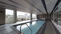This impressive indoor pool facility is designed with a clean, modern aesthetic that combines functionality with an elevated sense of luxury. At its centerpiece is a generously sized lap pool, finished in cool blue tones and framed by smooth tile surround decking that extends throughout the space. The pool's calm, reflective surface is complemented by stainless steel entry rails, lending a polished, professional quality to the environment.

The space is bathed in natural light, courtesy of expansive floor-to-ceiling windows that line the perimeter and open the interior to sweeping views of the surrounding cityscape. The structural ceiling features linear recessed detailing with subtle banding, contributing to the architectural sophistication of the room.

Along the windowed edge, a thoughtfully arranged collection of contemporary lounge chairs and small side tables provides a comfortable retreat for relaxation before or after a swim. The seating area benefits directly from the abundant natural light and the urban panorama beyond the glass.

The overall ambiance is calm, refined, and purposefully designed — a space that balances the practical demands of an aquatic facility with the refined character of an upscale amenity. The combination of tile work, modern furnishings, and architectural detailing creates an environment that is both inviting and visually striking.