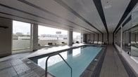 This impressive indoor pool facility is designed with a clean, modern aesthetic that combines functionality with an elevated sense of luxury. At its centerpiece is a generously sized lap pool, finished in cool blue tones and framed by smooth tile surround decking that extends throughout the space. The pool's calm, reflective surface is complemented by stainless steel entry rails, lending a polished, professional quality to the environment.

The space is bathed in natural light, courtesy of expansive floor-to-ceiling windows that line the perimeter and open the interior to sweeping views of the surrounding cityscape. The structural ceiling features linear recessed detailing with subtle banding, contributing to the architectural sophistication of the room.

Along the windowed edge, a thoughtfully arranged collection of contemporary lounge chairs and small side tables provides a comfortable retreat for relaxation before or after a swim. The seating area benefits directly from the abundant natural light and the urban panorama beyond the glass.

The overall ambiance is calm, refined, and purposefully designed — a space that balances the practical demands of an aquatic facility with the refined character of an upscale amenity. The combination of tile work, modern furnishings, and architectural detailing creates an environment that is both inviting and visually striking.