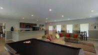 This single-floor space is designed with an open-concept layout that seamlessly blends recreation, relaxation, and functionality. The centerpiece of the main area is a full-size pool table, making it an ideal setting for social gatherings and leisure activities. Adjacent to the pool table is a generously sized sectional sofa accented with colorful throw pillows, providing ample comfortable seating for guests.

The kitchen area is positioned along one side of the space, featuring dark cabinetry, black appliances including a refrigerator and microwave, and a breakfast bar with counter seating — perfect for casual dining or entertaining. The open pass-through design keeps the kitchen visually connected to the main living and recreation area.

A dining area with a table and chairs is situated near the windows at the far end of the space, bathed in natural light from multiple windows fitted with white blinds. The neutral gray walls and recessed lighting throughout create a clean, well-lit atmosphere with a modern, casual feel.

Crown molding adds a subtle architectural detail, while the smooth ceilings and recessed can lighting ensure even illumination across the entire space. Overall, this layout is thoughtfully arranged to support a variety of social activities in one cohesive, welcoming environment.