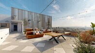 This expansive rooftop amenity level offers a thoughtfully designed outdoor experience with multiple distinct zones for relaxation, dining, and recreation. The deck is finished with large-format geometric tile throughout, lending a clean, contemporary aesthetic that ties the entire space together.

The main lounge area features substantial warm-toned wood sectional seating accented with colorful throw pillows, arranged around a bold wood-and-metal coffee table. String lights are strung overhead, creating an inviting atmosphere for evening gatherings. A ping pong table serves as a central recreational feature in this zone.

A secondary lounge area offers teak-framed chairs with classic navy-and-white striped cushions, positioned along the glass perimeter railing to take full advantage of sweeping panoramic views of the surrounding cityscape and distant mountains.

The dining and entertaining zone is anchored by a steel pergola framing an al fresco dining table with rattan chairs, set atop warm wood decking. Adjacent to this area is a dedicated BBQ station, well-suited for outdoor cooking and entertaining. Raised planter beds with ornamental grasses and succulents add natural texture throughout.

Interior corridors connect the rooftop zones and are lined with curated potted plants set against crisp white walls, maintaining the building's modern design language. The overall ambiance is bright, airy, and well-appointed.