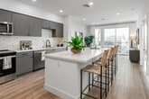 Located in the heart of Santa Ana, CA, this contemporary 2-bedroom, 1-bathroom residence offers a thoughtfully designed open-concept floor plan that maximizes space and natural light throughout. The modern kitchen is a standout feature, showcasing two-tone cabinetry in grey and muted blue, white quartz countertops, a large center island with bar seating, white subway tile backsplash, and a full suite of stainless steel appliances. Light wood-style flooring flows seamlessly from the kitchen into the bright living area, which opens through sliding glass doors to a private balcony overlooking the vibrant Santa Ana cityscape with views of palm trees and surrounding architecture. Both bedrooms are generously sized, each offering large windows that frame outdoor views and fill the rooms with natural light. The primary bedroom includes its own dedicated closet with built-in shelving and hanging rods for ample storage. The full bathroom features a clean, bright design with a tiled tub-shower combination, built-in niche shelving, a vanity with an illuminated mirror, and light tile flooring. Recessed lighting throughout the residence enhances the modern, airy ambiance of the entire space.