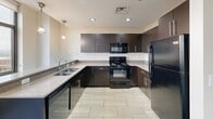 Located at 220 N Main St in downtown Davenport, Iowa, this two-bedroom, two-bathroom unit offers a well-designed single-floor layout with a thoughtful mix of modern finishes and functional spaces. The kitchen is a standout feature, equipped with rich dark espresso cabinetry, granite countertops, black appliances including a range, microwave, and refrigerator, and light-toned tile flooring. A peninsula counter provides additional workspace and a natural transition to the living area, complemented by pendant and recessed lighting overhead. Both bedrooms are carpeted in a bold, patterned blue textile and are brightened by windows offering views of the surrounding downtown cityscape, with warm wood-toned baseboards and doors adding a cohesive design element throughout. The primary bedroom includes a closet with wood-paneled doors. The two bathrooms feature clean tile flooring and dark cabinetry vanities; one includes a full tub-shower combination with sliding glass doors. An in-unit laundry room houses a stacked washer and dryer alongside a water heater, providing added convenience. A connecting hallway ties the rooms together, creating a practical and organized flow throughout the space.