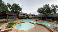Located at 3801 N Belt Line Rd in Irving, Texas, this outdoor community amenity space offers a well-appointed retreat designed for relaxation and outdoor living. The centerpiece is a generously sized swimming pool with clear turquoise water, complemented by a separate octagonal spa finished with natural stone coping, creating a resort-like atmosphere within the community.

Surrounding the pool, a concrete pool deck provides ample space for lounge chairs and seating arrangements, allowing residents to comfortably enjoy the outdoors. Stone-edged landscaping beds lined with river rock add a natural, polished aesthetic throughout the area, while mature trees provide shade and a sense of privacy.

A covered pergola with a warm wood-paneled ceiling serves as an outdoor gathering hub, sheltering a built-in stone grill station — ideal for outdoor cooking and entertaining. An adjacent wood deck extends the usable outdoor space, offering additional seating beneath the pergola's shade structure.

A separate covered pavilion near the pool provides further shaded seating and gathering space, reinforcing the communal nature of the amenity area. The grounds are well-maintained with manicured shrubs and green lawns that frame the space attractively. Together, these features create a cohesive, inviting outdoor environment well-suited for both quiet relaxation and social gatherings.