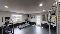 This single-floor fitness center is designed to support a well-rounded workout routine in a clean, functional environment. The space is finished with dark rubber flooring throughout, providing a durable and slip-resistant surface ideal for exercise. Neutral gray walls with a contrasting dark lower accent band give the room a modern, professional aesthetic, while recessed lighting ensures consistent illumination across the entire floor plan.

The cardio area is equipped with a Matrix treadmill and an elliptical cross-trainer, positioned near a large picture window that fills the space with natural light and offers views of the surrounding outdoor area. A wall-mounted flat-screen television is conveniently located nearby for entertainment during workouts.

The strength training section features a Hoist cable machine, a free-weight rack with a selection of dumbbells, and an adjustable workout bench. Full-length mirrors line the wall in this area, enhancing the sense of space and allowing users to monitor their form during exercises.

A dedicated stretch and functional training zone includes exercise balls, foam rollers, and additional floor space for flexibility and bodyweight work. Large sliding doors and a glass entry door connect this area to the outdoors, providing excellent ventilation and additional natural light. Posted fitness center rules and instructional signage complete this well-organized and accessible facility.